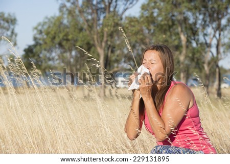 Portrait attractive mature woman suffering from seasonal hayfever allergy, sitting in field of blossoming wheat grass sneezing into handkerchief tissue, blurred outdoor background and copy space.