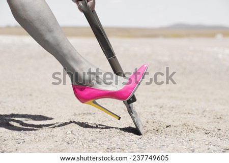 Concept filtered image of female leg in pink high heel stiletto shoe and one hand on shovel, digging in remote sandy desert hole in the dirt, blurred background and copy space.