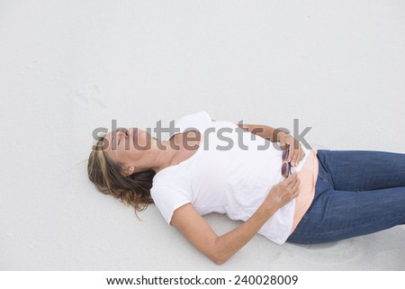 Portrait relaxed attractive mature woman lying thoughtful and sleepy dreaming with closed eyes on sandy beach, with sunglasses, white shirt and jeans, bright sand background and copy space.