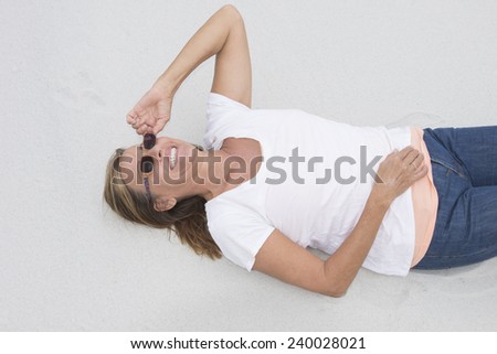 Portrait laid back attractive mature woman with sunglasses  lying happy smiling on sandy beach, white shirt and jeans, bright background and copy space.