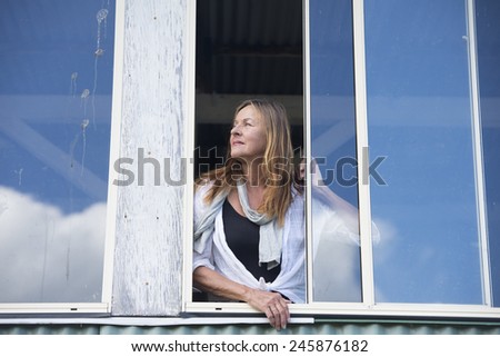 Portrait attractive mature woman standing at window watching, looking outside with serious, concerned expression, copy space.