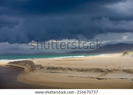 Dark dramatic storm cloud formation approaching ocean along bright sunny remote beach mountains in blurred background, copy space.