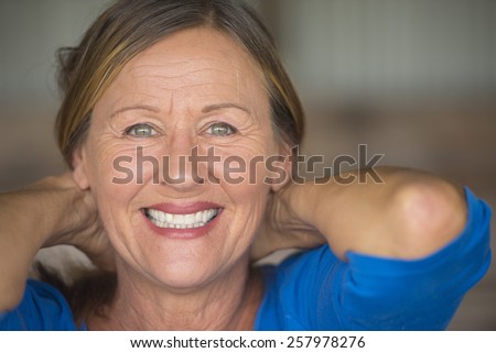 Portrait attractive mature woman with friendly positive confident happy smiling, hands behind neck, blurred background.