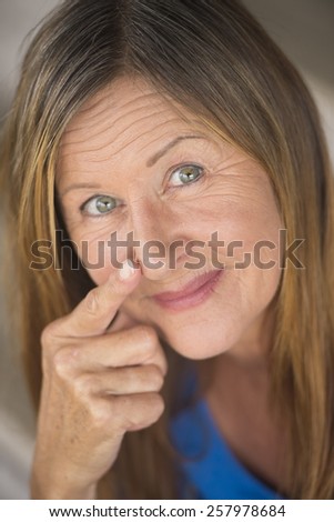 Portrait attractive mature woman with serious happy confident facial expression, upward look smile, finger pointing to nose, blurred background.