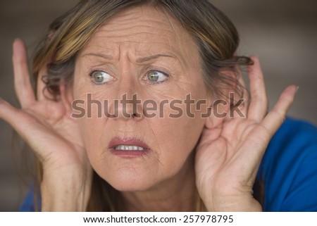 Portrait attractive mature woman with hands at ears listening with worried and anxious facial expression to loud noise or sound, blurred background.