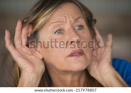 Portrait attractive mature woman with hands at ears listening with curious, interested and surprised facial expression to loud noise or sound, blurred background.