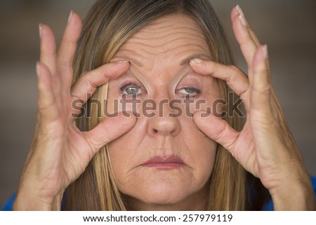 Portrait of attractive mature woman with tired and stressed facial expression, fingers keep eyes open, suffering migraine and headache, blurred background.