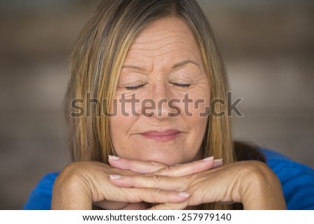 Portrait attractive mature woman with happy relaxed smiling facial expression, chin on hands, confident and peaceful daydreaming with closed eyes, blurred background.