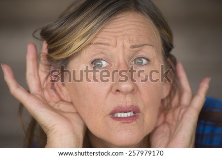 Portrait attractive mature woman with hands at ears listening with curious, interested and anxious facial expression to loud noise or sound, blurred background.