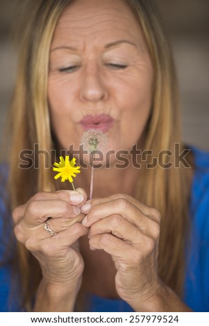 Portrait of attractive mature woman in blurred background blowing dandelion flower in hand, happy, friendly.