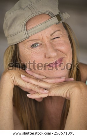 Portrait attractive fit active mature woman wearing green sporty top and cap, friendly funny, chin resting on hands, one closed eye, blurred background.