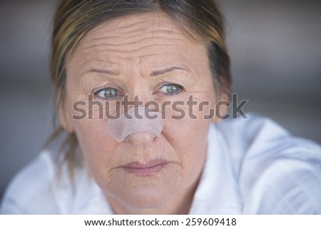 Portrait attractive mature woman unhappy stressed in pain with band aid on nose, blurred background, copy space.