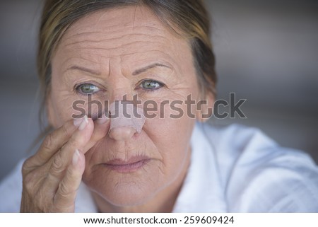 Portrait attractive mature woman stressed in pain with band aid on nose and unhappy expression, blurred background.