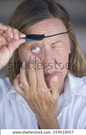Portrait injured attractive mature woman lifting eye patch worn as protection, blurred background.