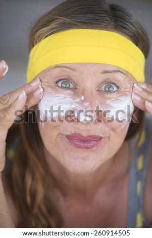 Portrait sporty fit active attractive mature woman with protective sunscreen skin care creme and moisturiser lotion on joyful smiling face, yellow headband, blurred background.