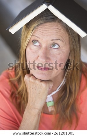 Portrait attractive mature woman with book on head, thoughtful look, waiting for inspiration and idea, blurred background.