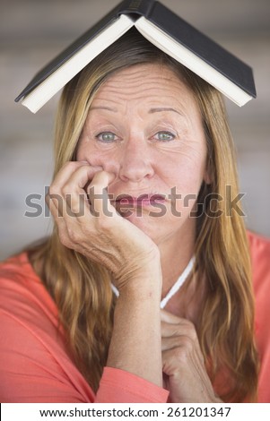 Portrait attractive mature woman with book on head, worried look, waiting for inspiration and idea, blurred background.