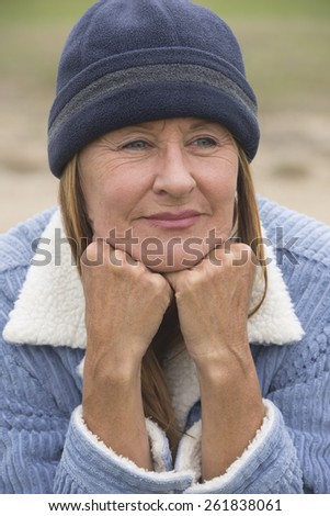 Portrait thoughtful attractive mature woman outdoor, wearing warm bonnet and wool jacket, resting relaxed chin on hands blurred green background.