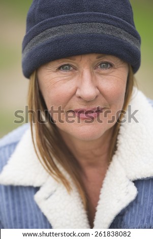 Portrait confident attractive mature woman outdoor, wearing warm blue bonnet and wool jacket, blurred green background.