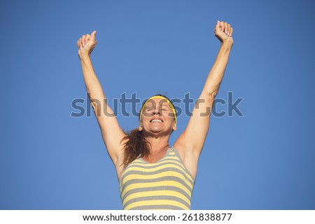 Portrait successful active attractive mature woman in victory pose, sporty, healthy, fit, arms up, closed eyes, blue sky background, copy space.