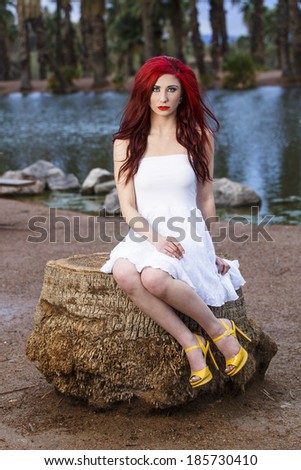 Young redhead in a white dress sitting on a rock by a lake
