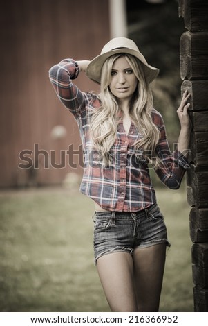 Southern and country blond girl posing in western wear with a hat outdoors by an old barn