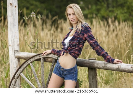 Young cowgirl in denim shorts and red color flannel top posing next to a fence and wagon wheel
