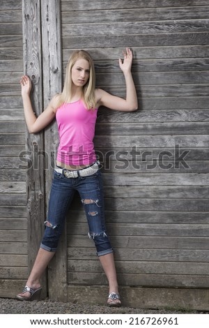 Young blond girl in pink top and ripped jeans posing with both arms up next to a wooden wall