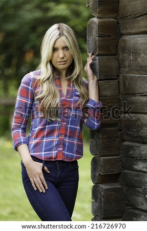 Young blond girl with straight hair posing next to a log cabin with red and blue flannel top and tight jeans