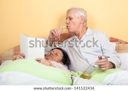 Funny senior couple relaxing in bed with alcohol and book