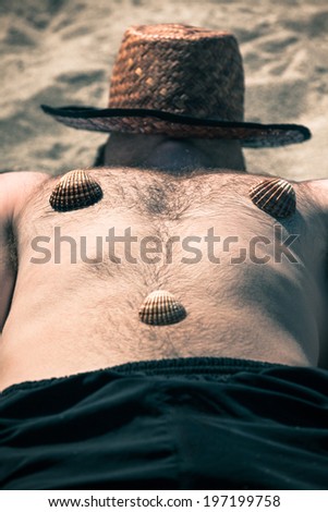 Funny shirtless man with hat and seashells on his body sleeping on the beach.