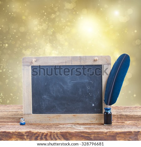 Blue  feather pen in inkwell on  aged wooden table with empty blackboard on glowing christmas background