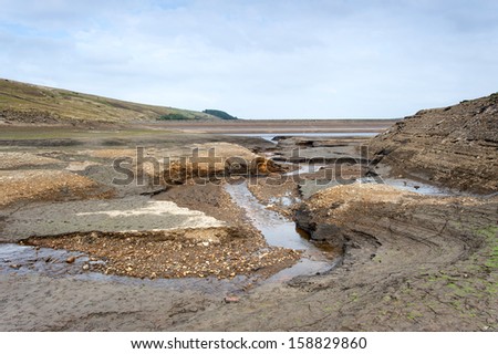 a muddy dried up stream leading the eye towards a yorkshire reservoir with falling water supply levels
