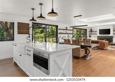 White and Bright Kitchen, Dining, and Living Room in New Luxury Home with Open Concept Floor Plan. Kitchen Features Island with Microwave and Farmhouse Sink. Shows View of Trees Through Windows.