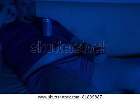 Middle-age, over-weight man watching TV alone in the dark, holding a can of beer on his big belly.