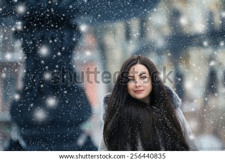 Beautiful girl with long dark hair in a beautiful coat and a long dress against a background of a winter landscape of the city, showfall
