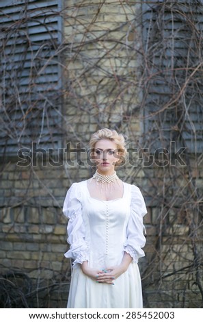 Beautiful blonde in an elegant white dress with a fan in solitude near the old house, fantastic atmosphere