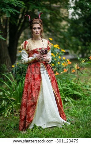 Beautiful girl with a demonic horns in medieval red dress on a background of nature with dry roses in their hands