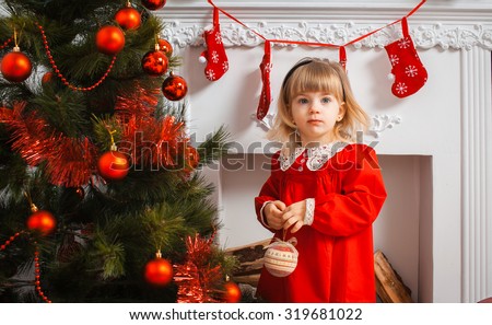 Cute little girl in a red dress against a white fireplace with red socks and Christmas tree, New Year\'s Day