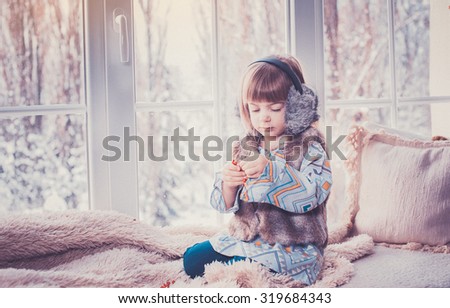 A cute little girl in warm clothes sits on a window sill, winter, snow