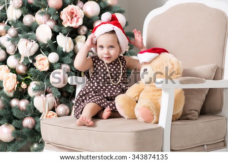 Cute little girl at the age of just over one year in the red cap of Santa Claus trying to teddy bear another red Santa hat against the backdrop of a beautiful Christmas tree