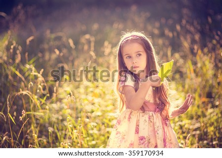 Cute little girl in a beautiful dress plays in the field at sunset with a large leaf of the tree