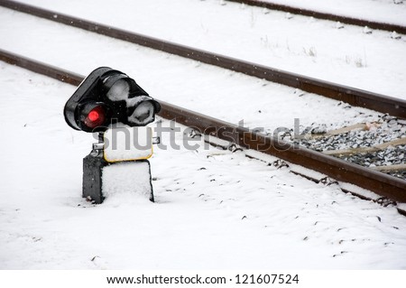 A red traffic light signal near a railroad track covered with snow