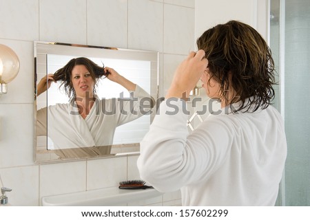 A woman is looking in the mirror of the bathroom and having a \'bad hair day\'.