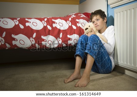 A young boy is sitting sad and depressed against his bed in his bedroom