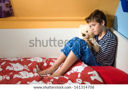 A young boy is sitting sad and depressed on his bed in his bedroom