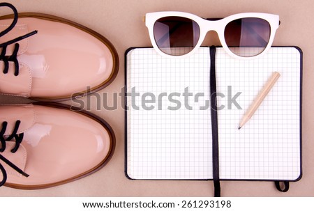 Shiny beige shoes on low heels, white sunglasses, wooden pencil and small black paper notebook. Beige background