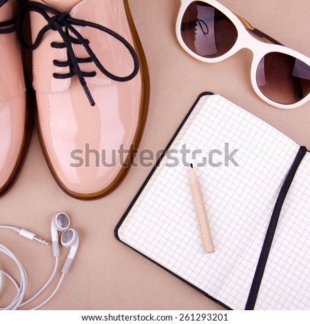 Shiny beige shoes on low heels, white sunglasses, earphones, wooden pencil and small black paper notebook. Beige background
