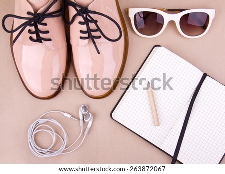 Shiny beige shoes on low heels, white sunglasses, earphones, wooden pencil and small black paper notebook. Beige background