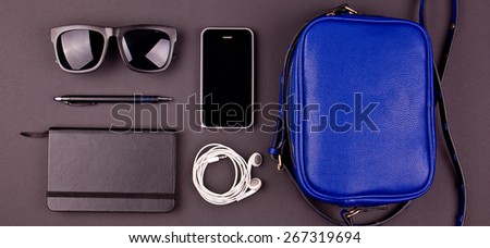 Still life of random objects of modern human. Handbag, cellphone, sunglasses, earphones, paper notepad, pen. Dark gray background. Aerial view. Concept.